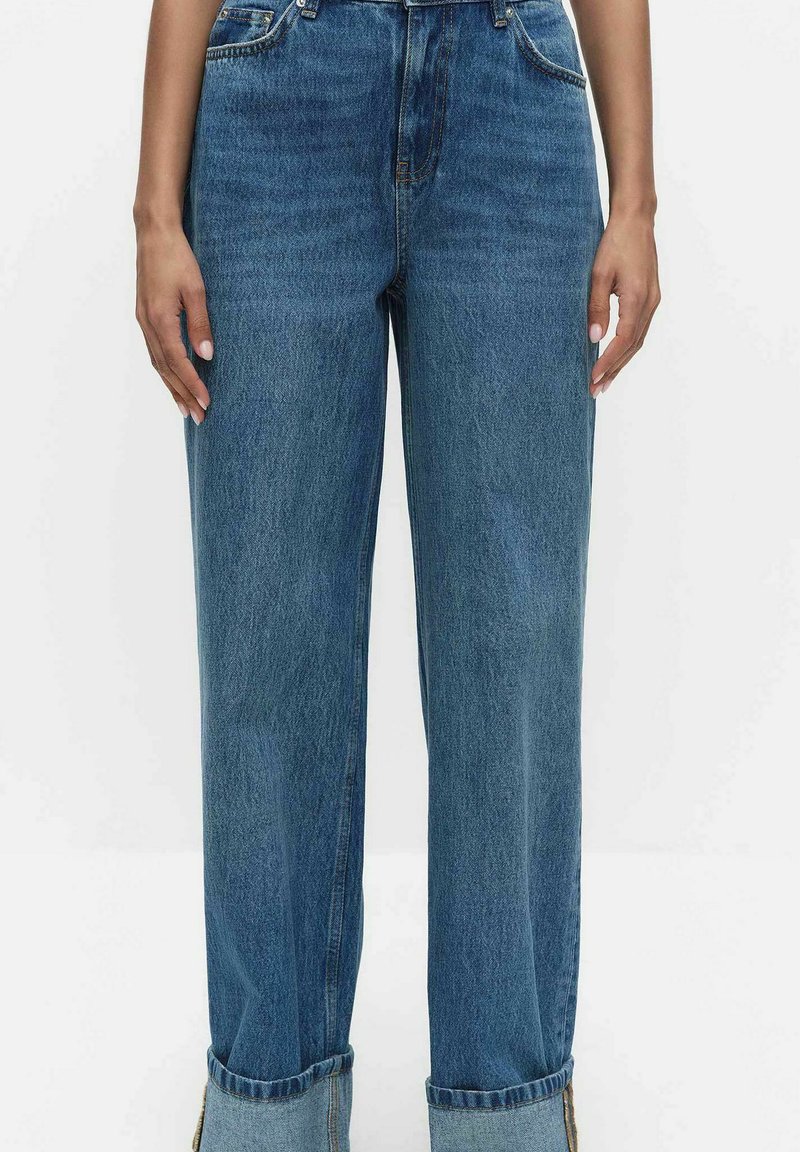 Kvinde iført blå jeans med høj talje og løstsiddende pasform med op rullede kanter, stående foran en ensartet hvid baggrund.