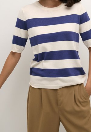 Personne portant un haut à manches courtes avec de larges rayures horizontales bleues et blanches et un pantalon ample beige, une main dans la poche.