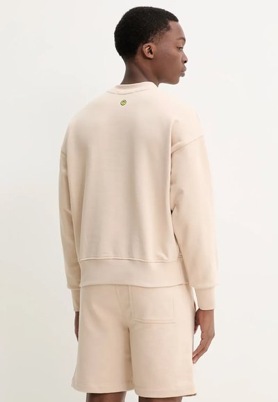Felpa beige con una vestibilità comoda, maniche a costine e orlo a coste. Un piccolo logo di smiley giallo è posizionato sul retro del collo. Abbinata a pantaloni corti coordinati.