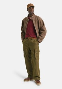 Mann mit braunem Hut, strukturiertem braunem Jackett, rotem Cable-Knit-Pullover, olivfarbenen Cargo-Hosen mit Taschen, geflochtenem Gürtel und braunen Schuhen, der mit den Händen in den Taschen dasteht.