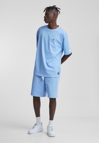 T-shirt bleu ciel à manches courtes avec logo brodé, associé à un short assorti. Des baskets blanches et des chaussettes texturées complètent la tenue.