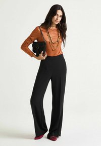 Haut orange à manches longues avec des pois, pantalon noir à jambes larges, sac à main noir orné de perles, colliers superposés et chaussures rouges embellies.