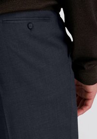 Close-up van een persoon in donkerblauwe broek met een knoopzak aan de achterkant en een donker langemouwenshirt dat zichtbaar is op hun arm.