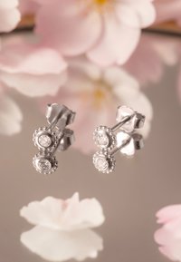 Silverörhängen med två cirkulära inställningar, var och en prydd med en klar ädelsten och intrikata pärldetaljer. Reflekterade på en spegelyta.