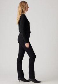 Top negro de manga larga combinado con jeans negros de talle alto y botas de tacón negro de punta. Textura suave, diseño ajustado, vista lateral.