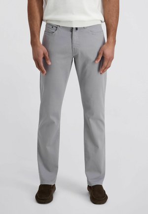 Man draagt lichtgrijze rechte broek, wit overhemd en donkerbruine schoenen, staand tegen een effen grijze achtergrond.