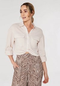 Hellbeige Hemd mit Knöpfen und Struktur sowie zwei Brusttaschen, kombiniert mit braun-weiß gestreiften Weitbundhosen.
