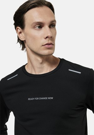 Ung mand med kort brunt hår bærer en sort skjorte med reflekterende skulderstriber og teksten "KLAR TIL FORANDRING NU" på brystet.