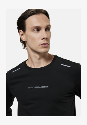 Ung mand med kort brunt hår bærer en sort skjorte med reflekterende skulderstriber og teksten "KLAR TIL FORANDRING NU" på brystet.