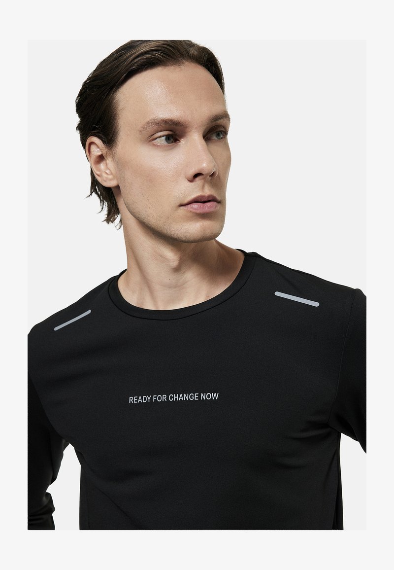 Młody mężczyzna z krótkimi brązowymi włosami nosi czarną koszulkę z odblaskowymi paskami na ramionach oraz napisem „READY FOR CHANGE NOW” na piersi.