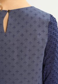 Mörkblå kläde med textur som har broderade blommönster, detaljerad nyckelhålsringning och texturerade ärmar.