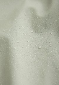 Ūdeni atgrūdošs audums gaiši bēšā krāsā ar daudzām izteiktām ūdens pilītēm uz gludas, teksturētas virsmas.