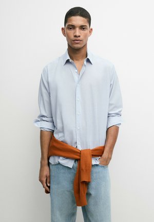 Giovane uomo che indossa una camicia azzurro chiaro con bottoni e maniche arrotolate, jeans chiari e un maglione arancione legato in vita.