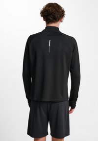 Zwart sportshirt met lange mouwen, van een textuurstof, met een opstaande kraag en een reflecterend logo op de rug, gecombineerd met zwarte shorts.
