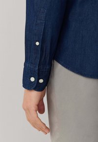 Koszula dżinsowa w ciemnoniebieskim odcieniu, z zapięciem na guziki przy mankietach oraz zaokrąglonym dołem. Widoczne szwy na końcach rękawów podkreślają detale. Miękka tekstura.