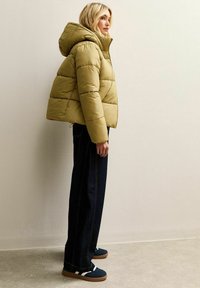 Chaqueta acolchada en color caqui con capucha, paneles texturizados y cierre de cremallera. Combinada con vaqueros oscuros de corte recto y zapatillas náuticas azul marino con rayas blancas.