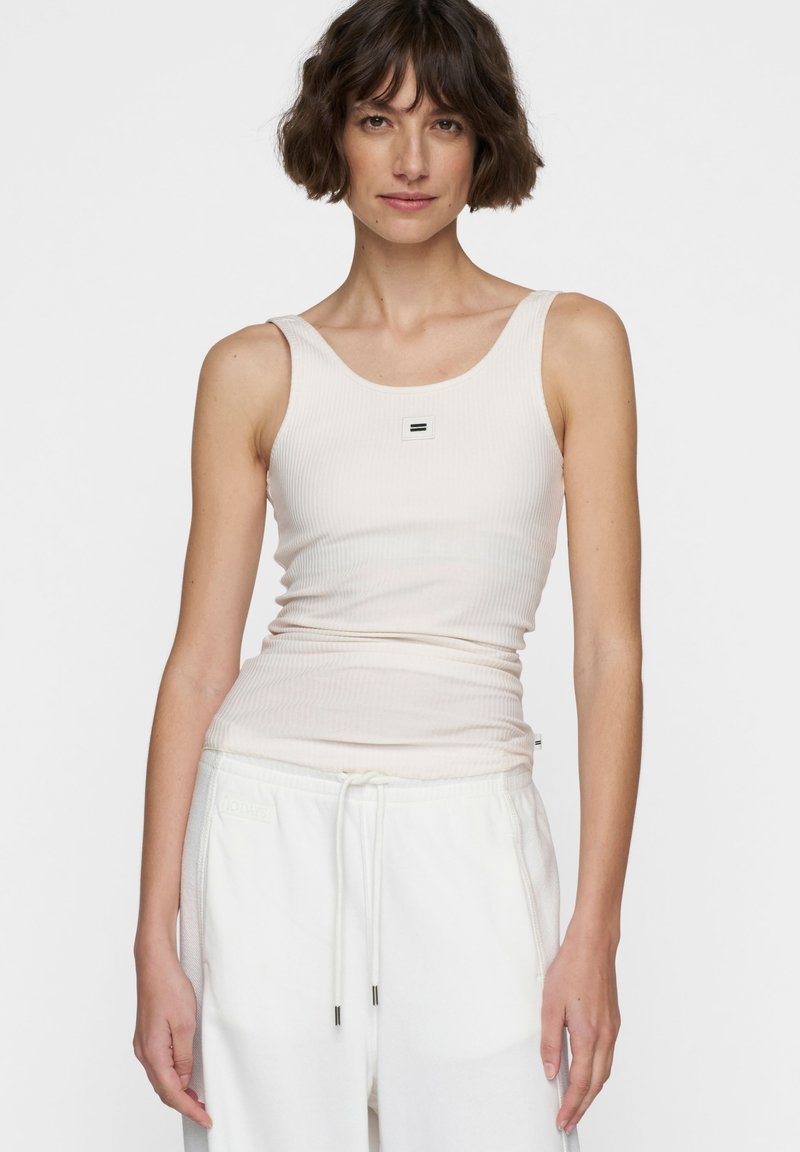 Canotta a coste beige chiaro con scollo rotondo e un piccolo logo quadrato nero al centro. Abbinata a pantaloni sportivi bianchi con coulisse.