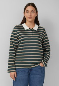 Frau mit langen braunen Haaren, die ein dunkelgrünes, braunes und weißes gestreiftes Langarmpolohemd und bluejeans trägt, mit der Hand in der Tasche.