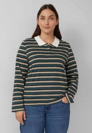 Vrouw met lang bruin haar, gekleed in een donkergroen, bruin en witte gestreepte lange mouwen polo shirt en blauwe jeans, met een hand in de zak.