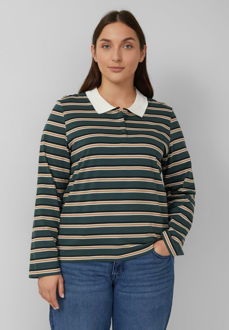 Frau mit langen braunen Haaren, die ein dunkelgrünes, braunes und weißes gestreiftes Langarmpolohemd und bluejeans trägt, mit der Hand in der Tasche.
