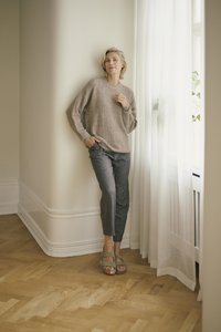 Light brown knitted sweater, grey tapered pants, and green sandals. Wood flooring and white curtains enhance the neutral color palette.