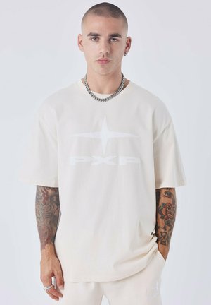 Camiseta oversized color crema en tejido suave con un gran logo blanco "PXP" y diseño de estrella en la parte frontal. Cuenta con mangas cortas y un corte relajado.