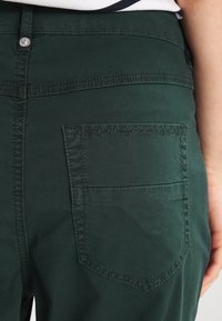 Pantalon en coton vert avec une poche arrière, des détails de couture et une taille ajustée. Le tissu a une texture lisse avec un léger éclat.