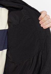 Giacca leggera nera con una superficie esterna liscia e chiusura frontale con zip. Presenta una tasca interna e dettagli di design minimalisti.
