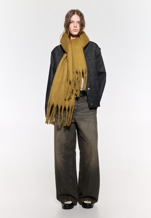 fular din lână khaki cu capete franjurate, purtat peste o jachetă din denim negru. Modelul asortează cu blugi gri deschis, cu picioare largi, și încălțăminte neagră.