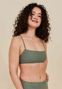 Donna con capelli ricci scuri che indossa un costume da bagno a due pezzi verde, sorridendo leggermente, su uno sfondo beige uniforme.