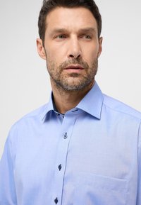 Camicetta azzurro chiaro con una texture liscia, collo button-down e bottoni blu navy. Presenta una tasca sul petto e un sottile motivo all'interno del colletto.