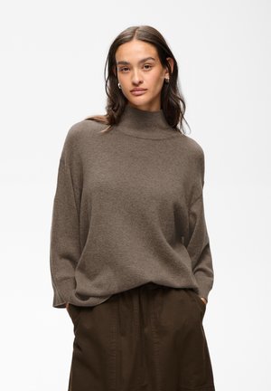 Jeune femme aux longs cheveux foncés portant un pull à col roulé marron ample et un pantalon marron foncé, les mains dans les poches.