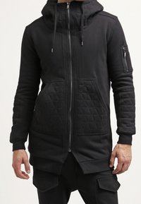 Felpa nera con un frontino trapuntato e cappuccio, dotata di zip completa, tasche laterali e una tasca con zip aggiuntiva sulla manica.