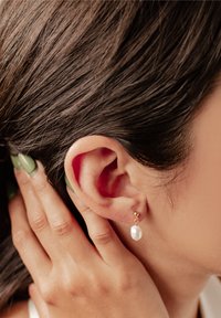 Złote kolczyki w kształcie kropli charakteryzują się eleganckim designem, z okrągłą złotą kulką i akcentem w postaci perły w kształcie łzy, delikatnie wisząc przy uchu.