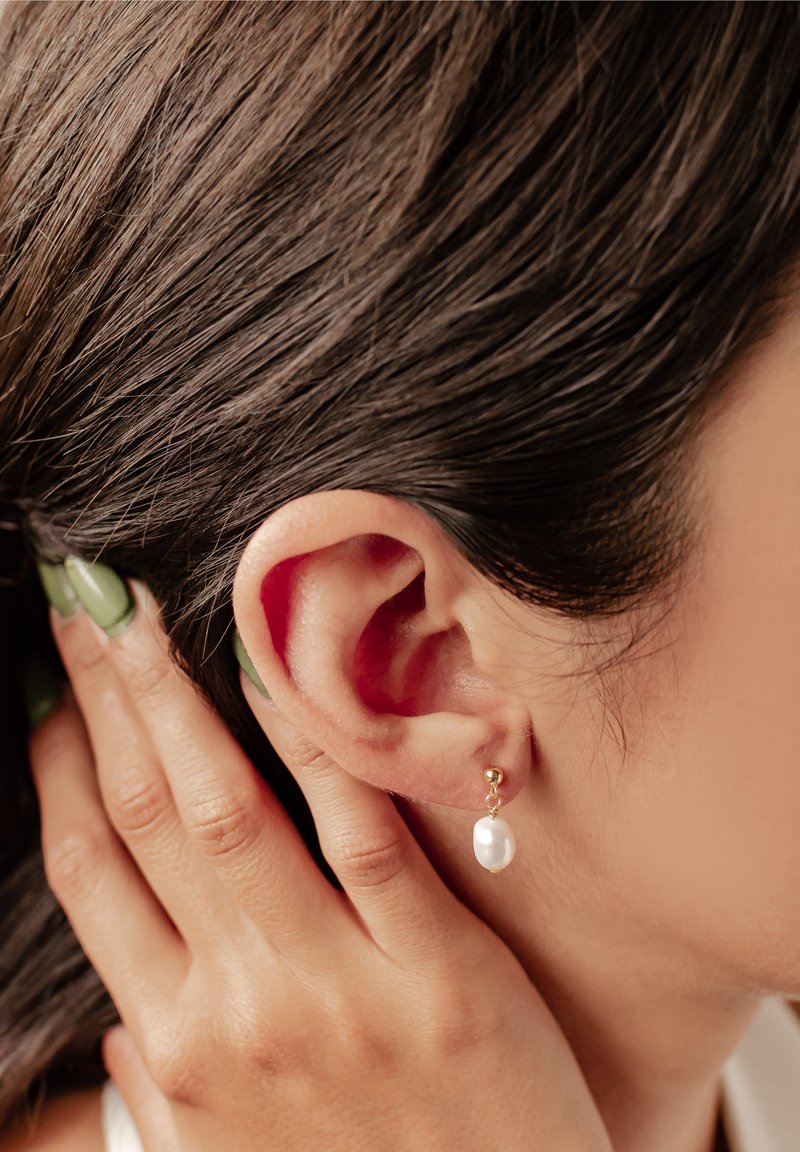 Złote kolczyki w kształcie kropli charakteryzują się eleganckim designem, z okrągłą złotą kulką i akcentem w postaci perły w kształcie łzy, delikatnie wisząc przy uchu.