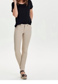 Mujer vestida con pantalones ajustados beige, top negro de manga corta y sandalias de cuña negras, de pie contra un fondo blanco liso.