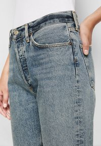 Gros plan sur une personne portant un jean en denim bleu clair taille haute avec une main posée dans la poche avant, associé à un haut blanc.