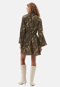Robe olive verte à manches courtes avec un motif paisley, taille cintrée et manches évasées ; le modèle porte des bottes blanches jusqu'aux genoux.