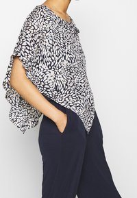 Femme portant un haut fluide à motifs noir et blanc avec des manches courtes et un pantalon bleu marine, se tenant avec une main dans la poche.