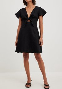 Femme portant une robe courte noire avec un nœud à l'avant et des manches volantées, associée à des talons ouverts noirs à brides, debout devant un fond uni.