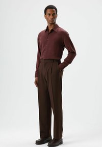 Bruin overhemd met knopen, gecombineerd met op maat gemaakte bruine broek. Het overhemd heeft een puntige kraag; de broek is geplooid en heeft zijzakken.