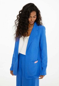 Modrá sako vyrobené z ľahkej tkaniny, s jednou kapsou a ružovým označením, so štruktúrovanými ramenami a mierne nadmernou veľkosťou.