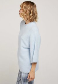 Pull en tricot bleu clair, confortable, avec un col rond et des manches trois-quarts. Texture douce et coupe décontractée. Pantalon à motif vichy visible.