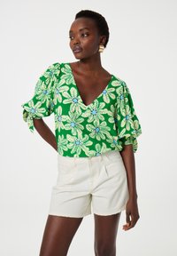 Groene bloemenblouse met korte, opbollende mouwen, voorzien van een V-hals en blauwe accenten. Gepaarde met crèmekleurige shorts.