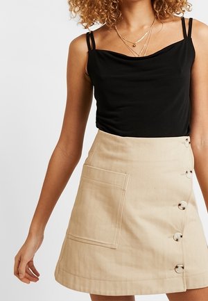 Frau trägt ein schwarzes ärmelloses Top mit dünnen Trägern und einen beigen, geknöpften Rock mit einer Fronttasche, gezeigt vom Hals bis zur Mitte des Oberschenkels.