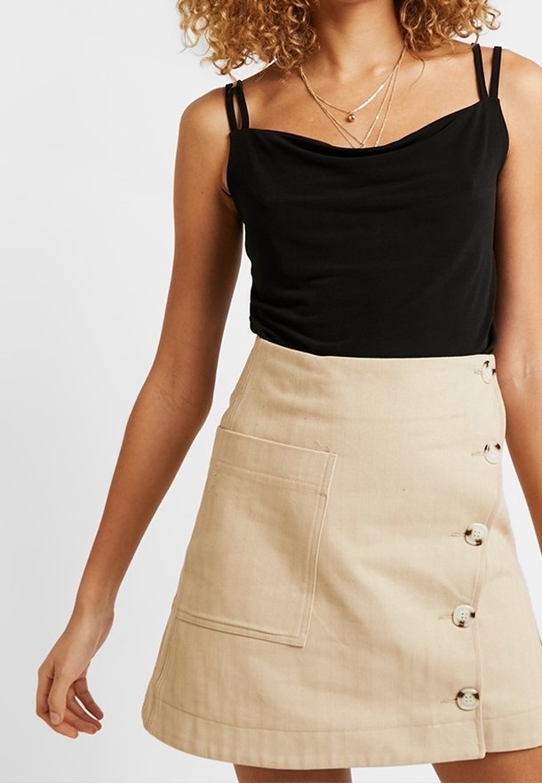 Femme portant un haut noir sans manches à fines bretelles et une jupe beige boutonnée avec une poche avant, montrée du cou jusqu'à mi-cuisse.