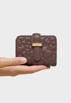 Mano che tiene un portafoglio in pelle marrone goffrata con una cinghia e il logo "TOUS" in tonalità oro su uno sfondo chiaro e semplice.