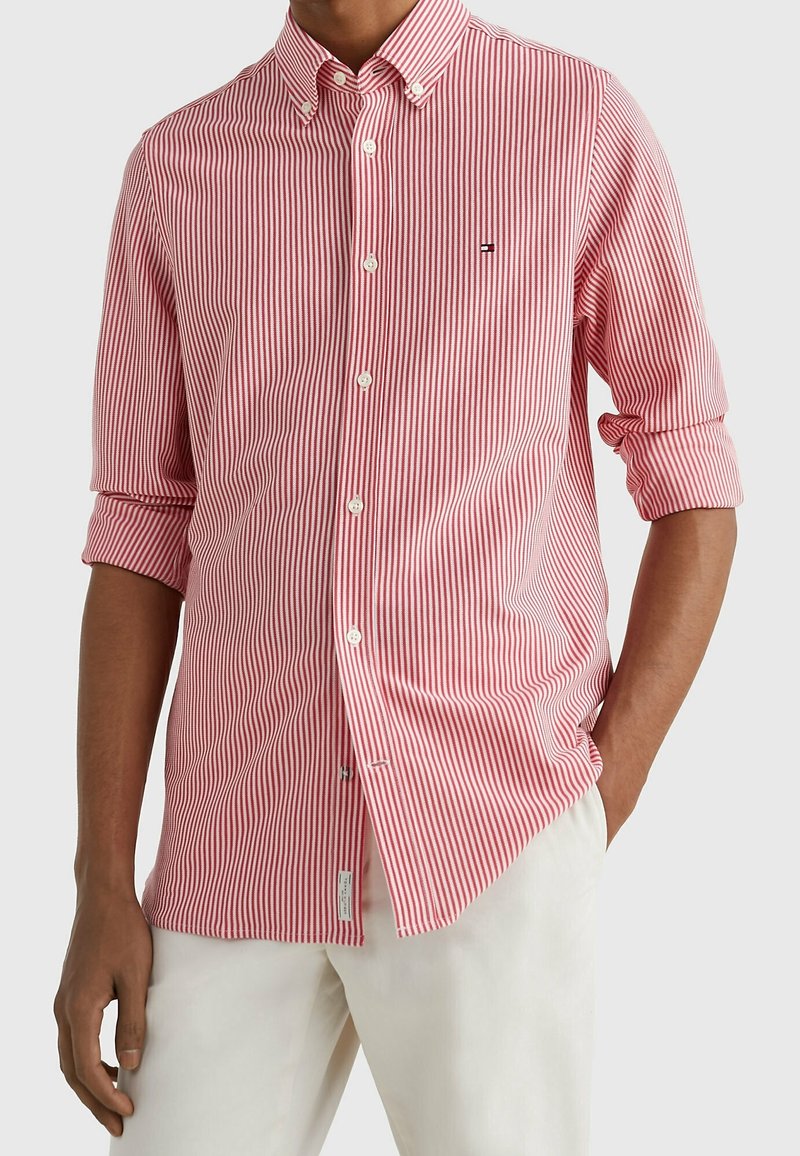 Homme portant une chemise à rayures verticales rouges et blanches à manches longues retroussées et un pantalon blanc, debout avec une main dans la poche.
