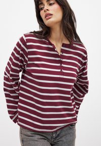 Burgunderfarbenes Langarmshirt mit weißen horizontalen Streifen, Rundhalsausschnitt und Knopfleiste mit drei Knöpfen. Weicher Stoff mit lockerer Passform.