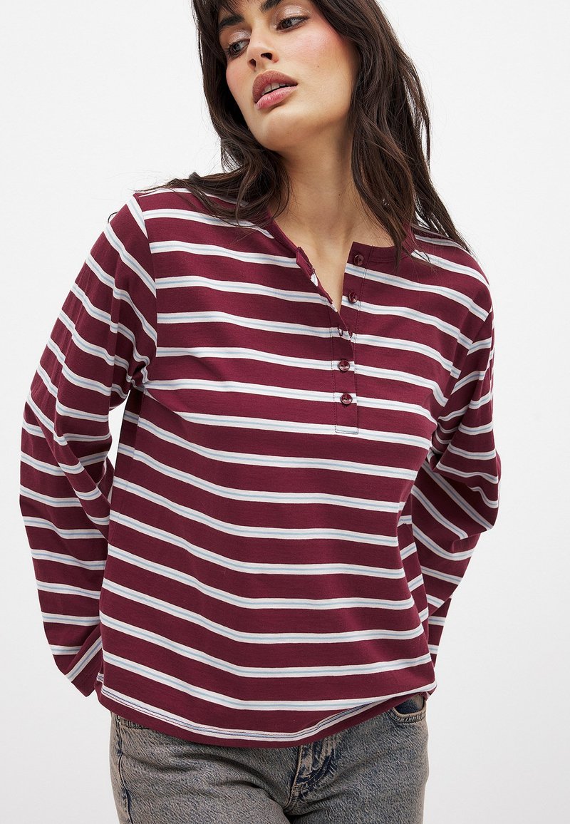 Burgunderfarbenes Langarmshirt mit weißen horizontalen Streifen, Rundhalsausschnitt und Knopfleiste mit drei Knöpfen. Weicher Stoff mit lockerer Passform.